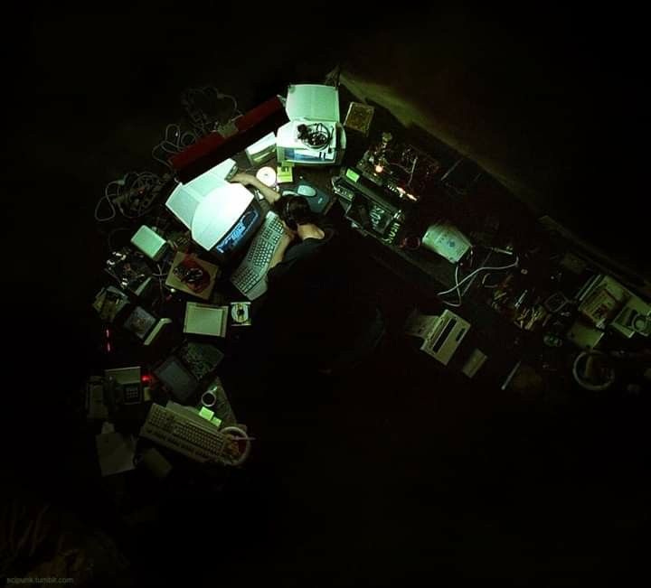 Overhead shot of a cluttered, dimly lit desk with old computer monitors glowing, cables and books scattered, and a person hunched over working late at night-evoking obsessive, Black Mirror vibes. 