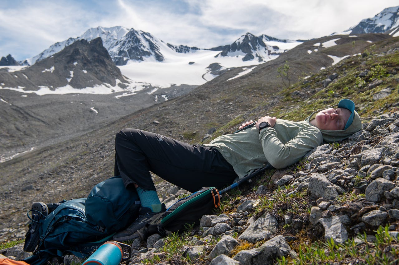 A hiker lies back on a tundra slope with eyes closed, head near her pack and hands folded on her chest, a cap shading her face. Behind her, a rubble-and-snow slope climbs to jagged snow-covered peaks under a partly cloudy sky.
