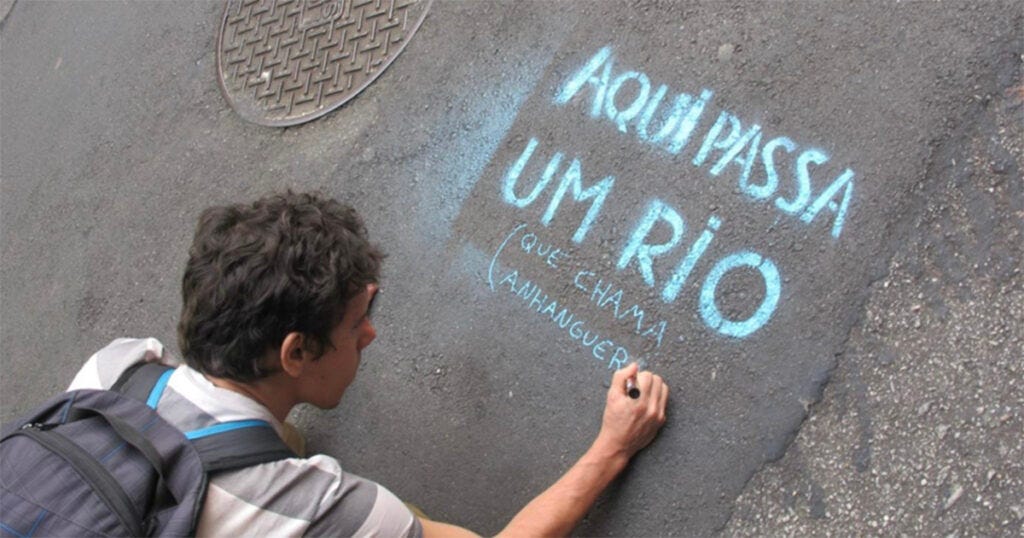 Montagem com quatro fotografias de intervenções do coletivo Aqui Passa um Rio, em São Paulo e uma do Bloco do Água Preta. As imagens mostram placas e pinturas com a frase “Aqui passa um rio” instaladas em diferentes pontos da cidade, como calçadas, ruas e estruturas urbanas, sinalizando a presença de rios ocultos pela urbanização. Montagem com quatro fotografias de intervenções do coletivo Aqui Passa um Rio, em São Paulo e uma do Bloco do Água Preta. As imagens mostram placas e pinturas com a frase “Aqui passa um rio” instaladas em diferentes pontos da cidade, como calçadas, ruas e estruturas urbanas, sinalizando a presença de rios ocultos pela urbanização.