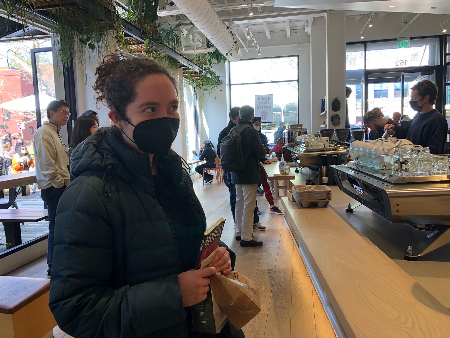 Shira in a mask and puffer coat looks out of the corner of her eye by a light wood counter