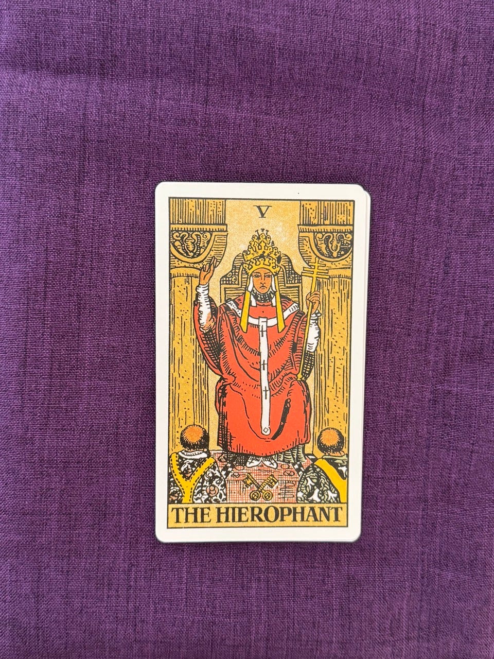 A robed religious figure sits facing forward on a stone throne between two gray pillars. He wears a red outer robe over white garments and a tall, ornate crown with three tiers. His right hand is raised in a gesture of blessing, with fingers positioned in a symbolic pose. In his left hand, he holds a staff topped with three horizontal bars. At his feet lie two crossed keys, one gold and one silver. Two kneeling figures in patterned robes face him with bowed heads. The background is plain and subdued, emphasizing a formal, ceremonial setting.