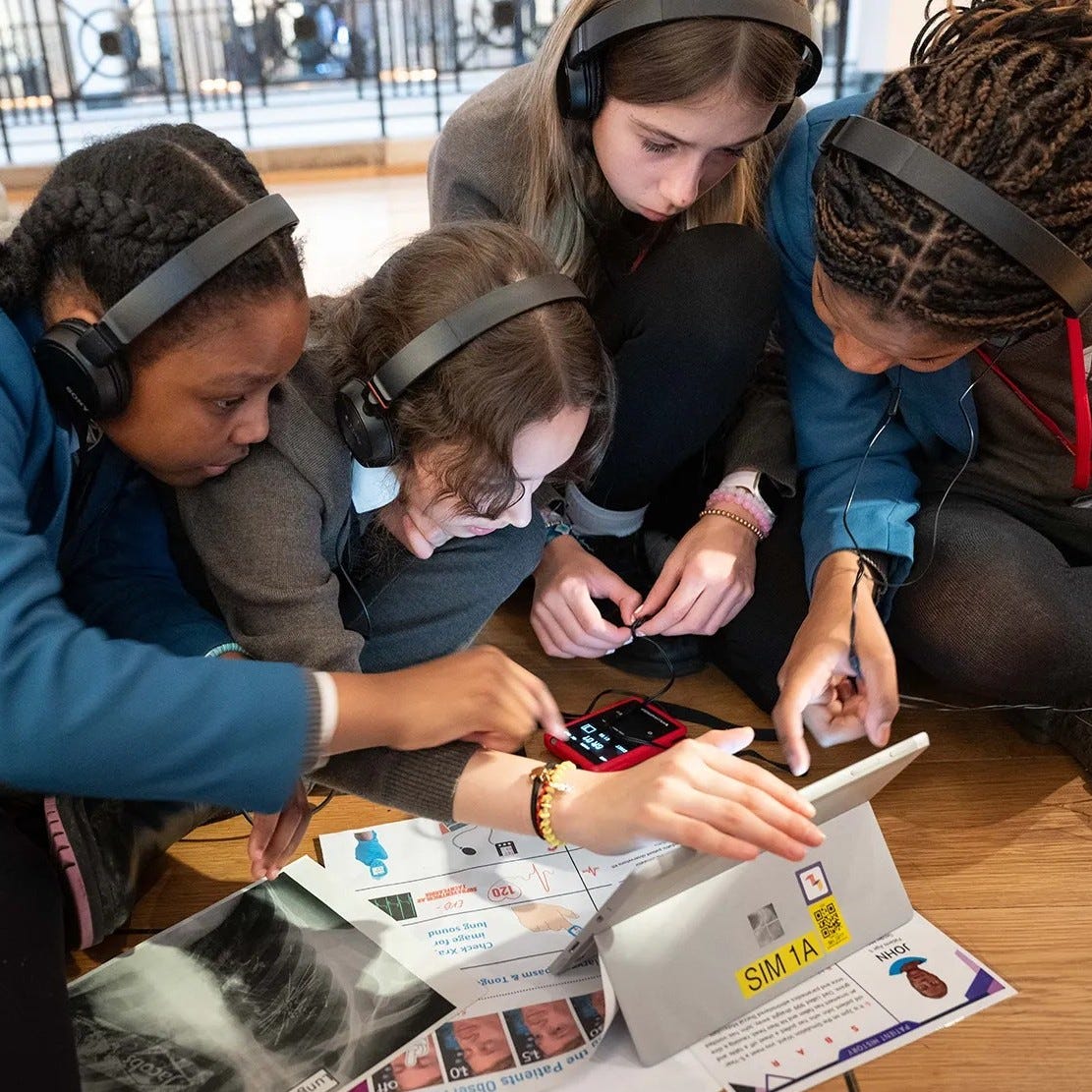 Four students wearing headphones collaborate around a tablet during a STEM learning activity in a museum setting.