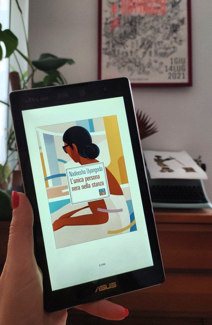 Una mano tiene un tablet. Sul tablet c'è la copertina di un libro: L'unica persona nera nella stanza. Rappresenta una persona nera che guarda fuori da una finestra. Tutta in colori pastello. Sullo sfondo, dietro al tablet, di vede un mobiletto marrone con sopra una pianta e una macchina da scrivere, sopra la quale c'è appoggiato un disegno. Appeso al muro, dietro al mobiletto, c'è un quadro con un disegno in bianco e nero. Il quadro ha una cornice rosa.