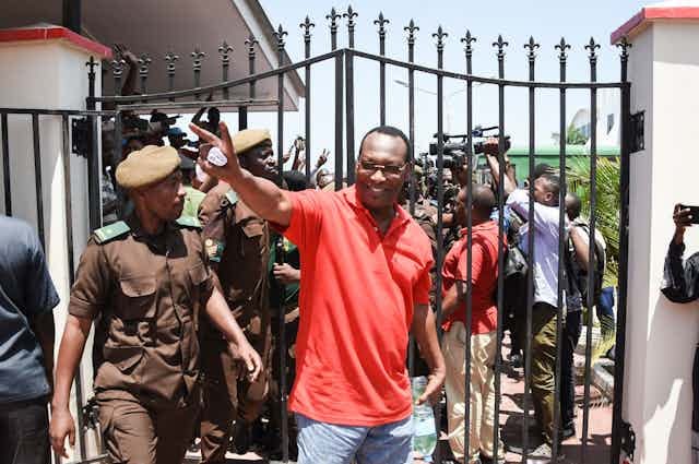 A man in a red shirt and wearing specs holds up two fingers in a wave while surrounded by soldiers in a brown uniform