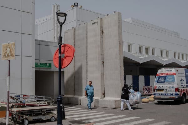 A person in scrubs walks outside of a concrete barrier near a large white building. An ambulance is parked as a person pushing a cart full of bags walks by.