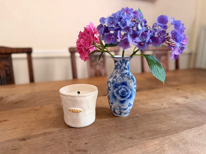 A woman walking through a furnished coach house sitting room; an open cupboard of folded white linens and towels; a woman in a straw hat clipping hydrangeas; a blue-and-white vase of pink and purple hydrangeas.