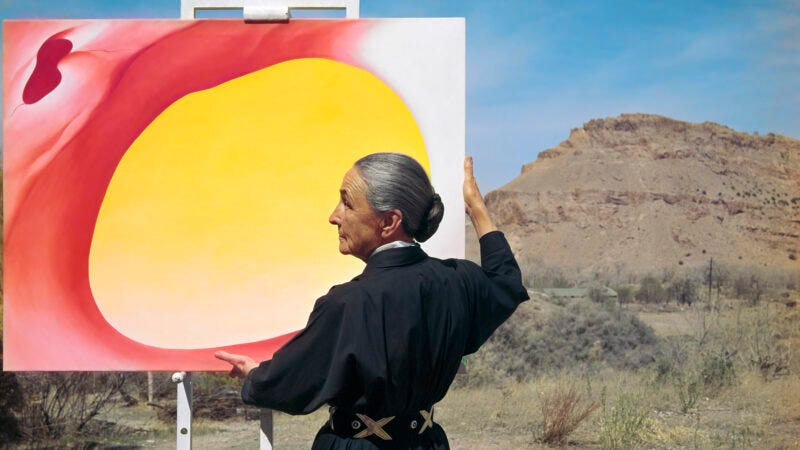Photograph of Georgia O'Keeffe holding a painting in front of a landscape of desert mountains
