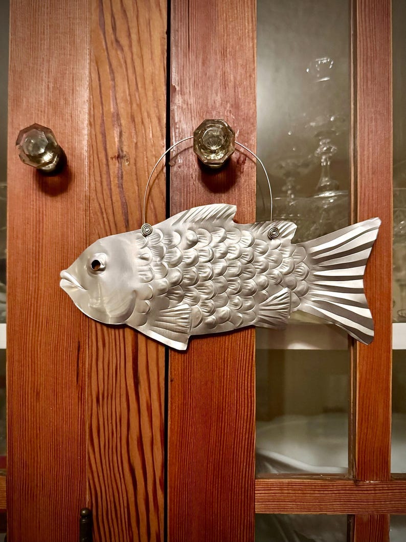 May include: A silver metal fish with a textured, scaly surface hangs from a wire loop on a wooden cabinet door. The fish has a slightly open mouth and a fin on its back. May include: A silver metal fish with a textured, scaly surface hangs from a wire loop on a wooden cabinet door. The fish has a slightly open mouth and a fin on its back.