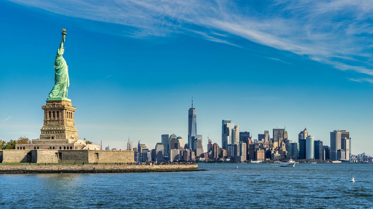 The Statue of Liberty on the left, the skyline of Manhattan in the middle and right of this mostly blue (water/sky) photograph.