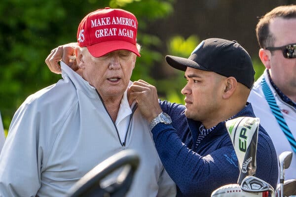 Donald Trump, wearing his trademark red cap, has his collar adjusted by Walt Nauta, wearing a black cap and blue pullover. 