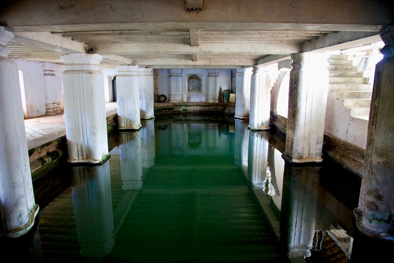 The flooded ground-floor bathing pool of the Queens' Bathing Pavilion in Kandy, with rows of white columns reflected in still green water. The flooded ground-floor bathing pool of the Queens' Bathing Pavilion in Kandy, with rows of white columns reflected in still green water.