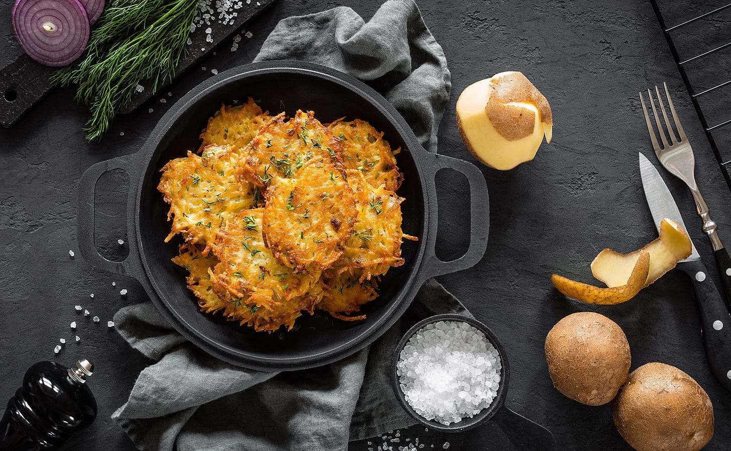 overhead view of golden potato pancakes in a black skillet on a table with a bowl of salt and a few whole potatoes