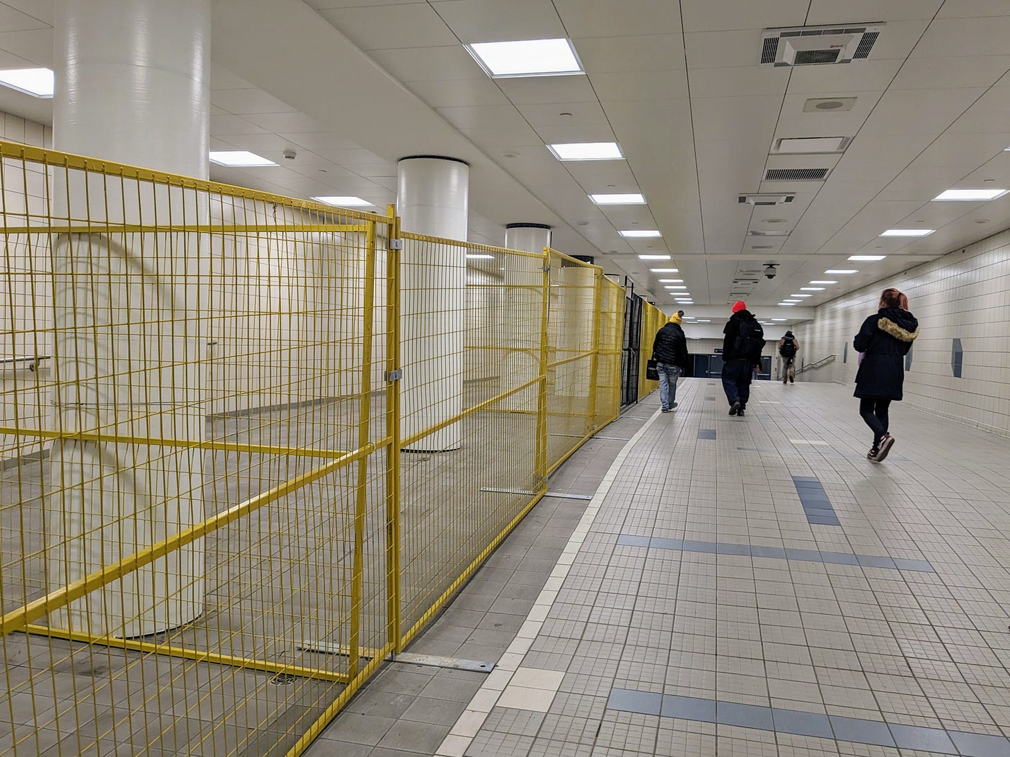 A transit tunnel in Edmonton is underground, showing people walking through alongside a yellow steel fence that closes off an open area in the tunnel, where on house people used to congregate and keep warm. Fluorescent lights beam overhead. The photo has a sense of coldness and Cavernous space A transit tunnel in Edmonton is underground, showing people walking through alongside a yellow steel fence that closes off an open area in the tunnel, where on house people used to congregate and keep warm. Fluorescent lights beam overhead. The photo has a sense of coldness and Cavernous space