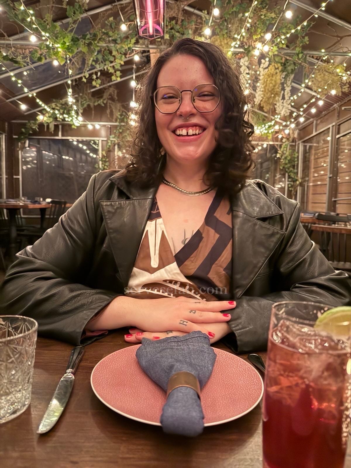 A beautiful fair skinned woman with shoulder length brown curly hair wears a leather jacket and smiles at you. You are her dinner date across the table. There are lights and greens strung all around the ceiling and walls, and heatlamps, as you are eating outside