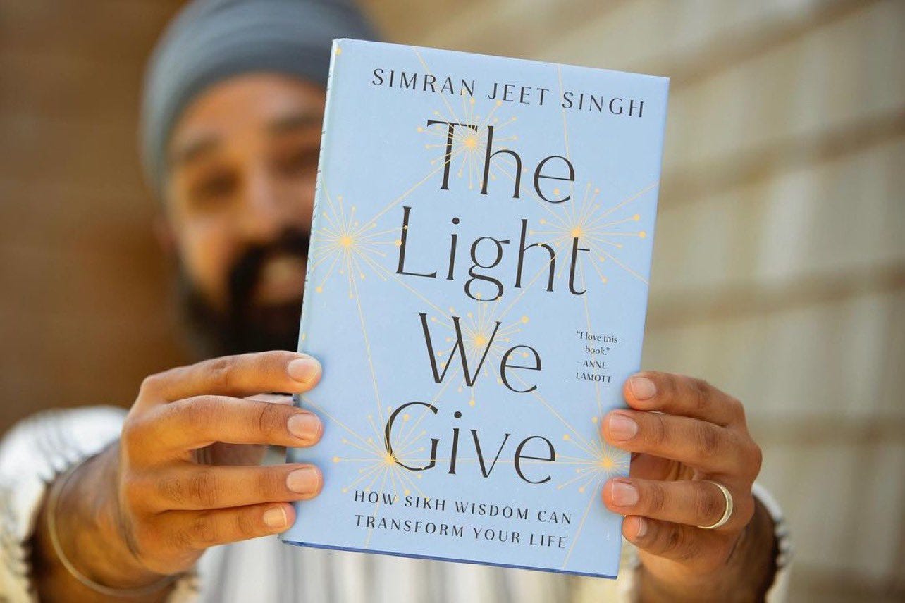 Simran is holding his book out in front of him. It's a light blue book, with yellow stars bursting out across the cover, with the title The Light We Give across the front in black font. 