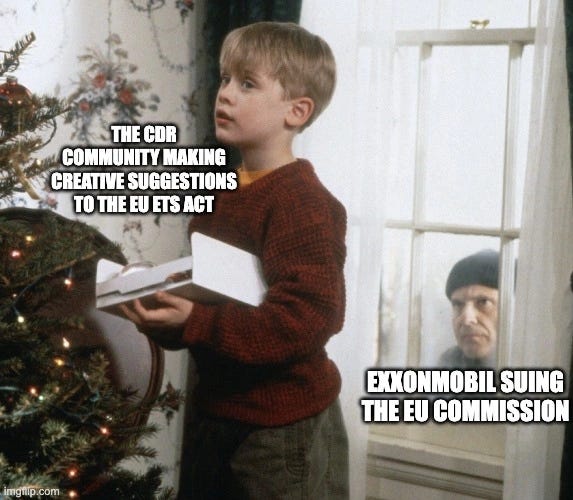 Still from Home Alone. Kevin, holding a box by a Christmas tree, is labelled "THE CDR COMMUNITY MAKING CREATIVE SUGGESTIONS TO THE EU ETS ACT". Through the window behind him an old man in a cap peers in, labelled "EXXONMOBIL SUING THE EU COMMISSION".