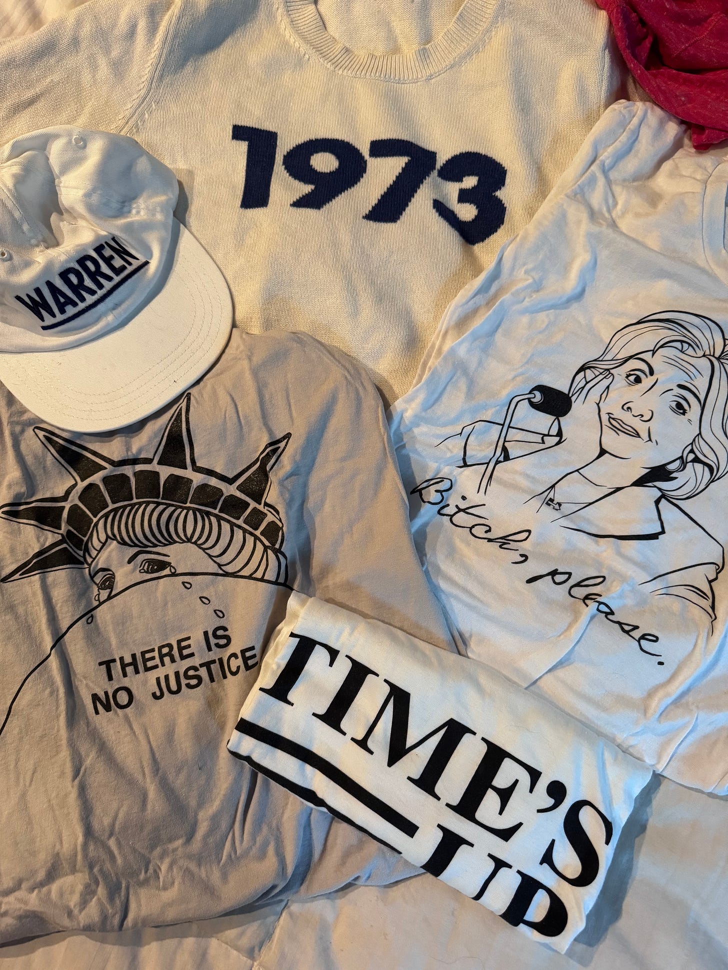Some of Amber’s political clothing laid out. There is the 1973 sweater; a t-shirt with a drawing of Hillary Clinton on it with the words “Bitch, please” underneath; a gray shirt with a crying Statue of Liberty with the words “There is no justice.” A white, flat brim baseball cap that says “Warren” in underlined black text; a white shirt with black text that reads “Time’s Up.”