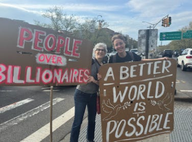 Photo of Julie and Kate at Freedom Friday protest, holding big signs that read "PEOPLE OVER BILLIONAIRES" and "A BETTER WORLD IS POSSIBLE."