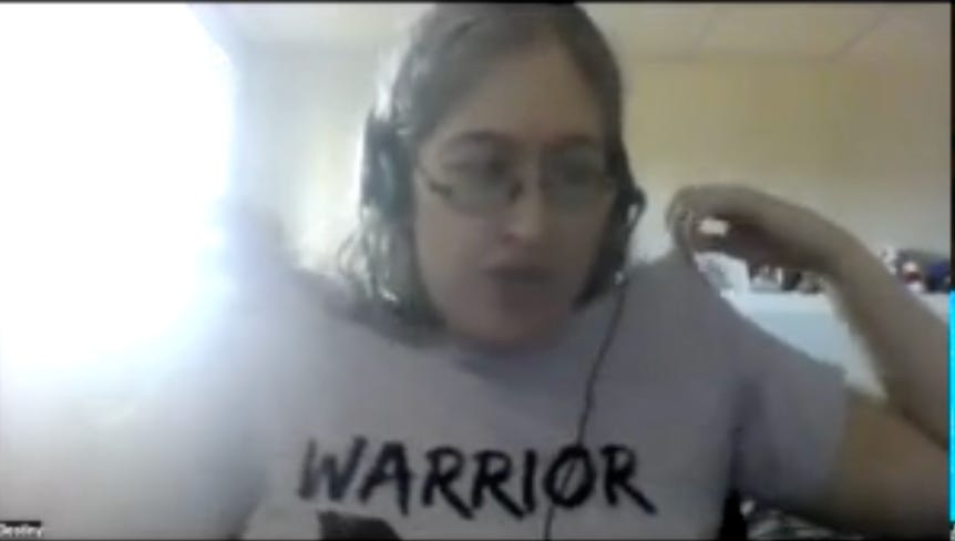 Destiny a woman with glasses and greying hair, wearing headphones on the computer, points to the shirt she is wearing that says WARRIOR Destiny a woman with glasses and greying hair, wearing headphones on the computer, points to the shirt she is wearing that says WARRIOR