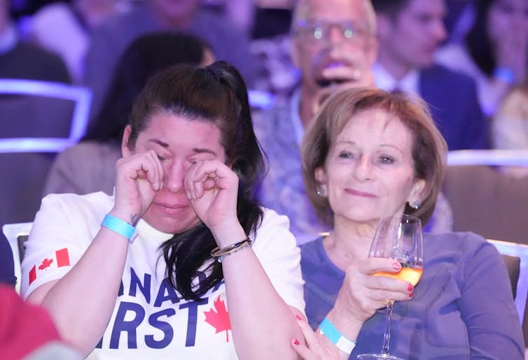 A woman with dark hair weeps while another woman sits next to her sipping a glass of wine. A woman with dark hair weeps while another woman sits next to her sipping a glass of wine.