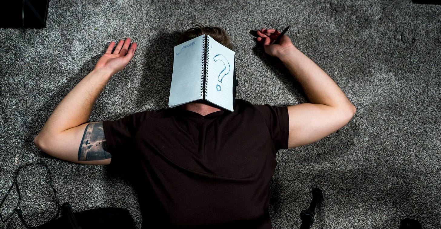 A man in a black t-shirt, tattoo visible under one sleeve on his biceps, lies flat on his back on grey carpeting, arms raised to either side, hands palm up. A spiral notebook lies open over his face, pages facing the viewer, a giant question mark penned onto the right side.