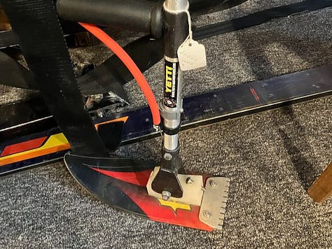 Left: A pinback button about 1" across sits in a museum display case. It was for the Jaycee Ski School and is red, white, and blue. Center: A sit-ski outrigger from the 1990s. It has an aluminum shaft and the very front tip of a ski attached to the end. Right: A white, black, and light blue button for the flying outriggers ski club. Their logo shows a pair of standing-ski outriggers, like crutches, with flags on them, waving in the wind..
