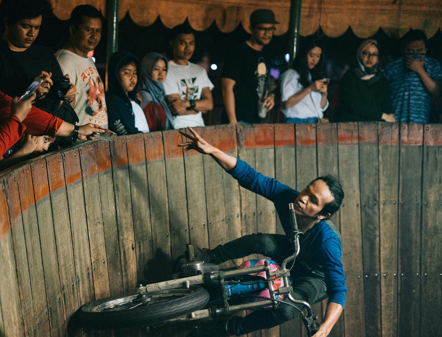 A daredevil motorcycle rider is halfway up a sheer wooden wall, reaching for a few bucks a spectator is giving him as a tip