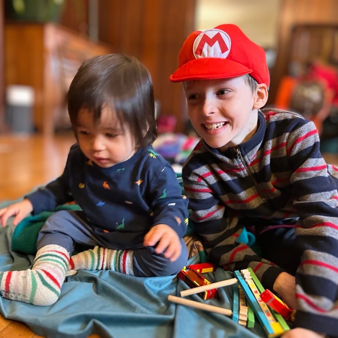 A young child in a red Mario cap and striped sweater grins while sitting on a soft teal blanket next to a toddler in a dark blue onesie decorated with colorful dinosaur patterns. Between them, a set of wooden percussion instruments is scattered, with the toddler focused on exploring them.