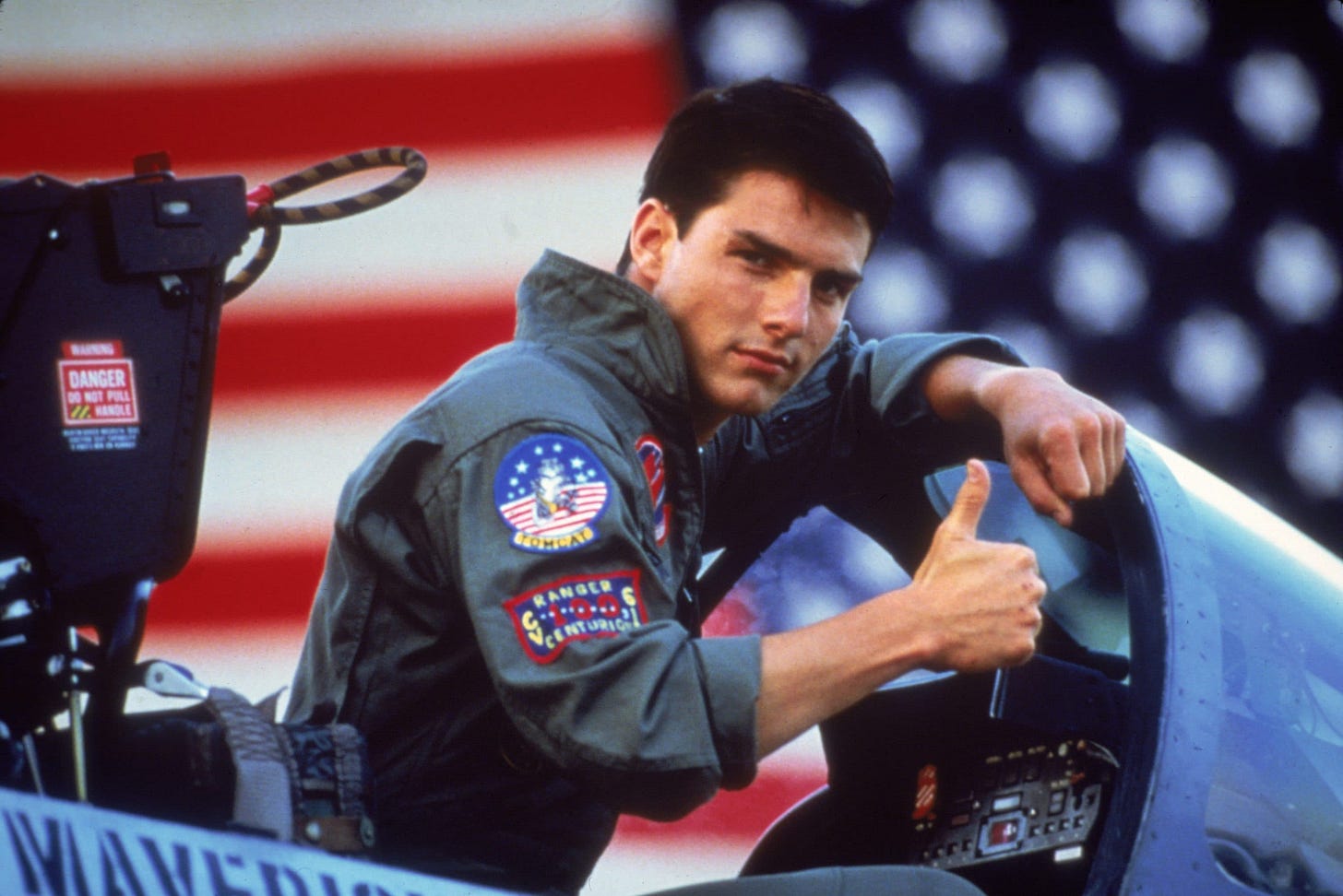 still from Top Gun; Tom Cruise giving a thumb's up in a cockpit, American flag in background still from Top Gun; Tom Cruise giving a thumb's up in a cockpit, American flag in background
