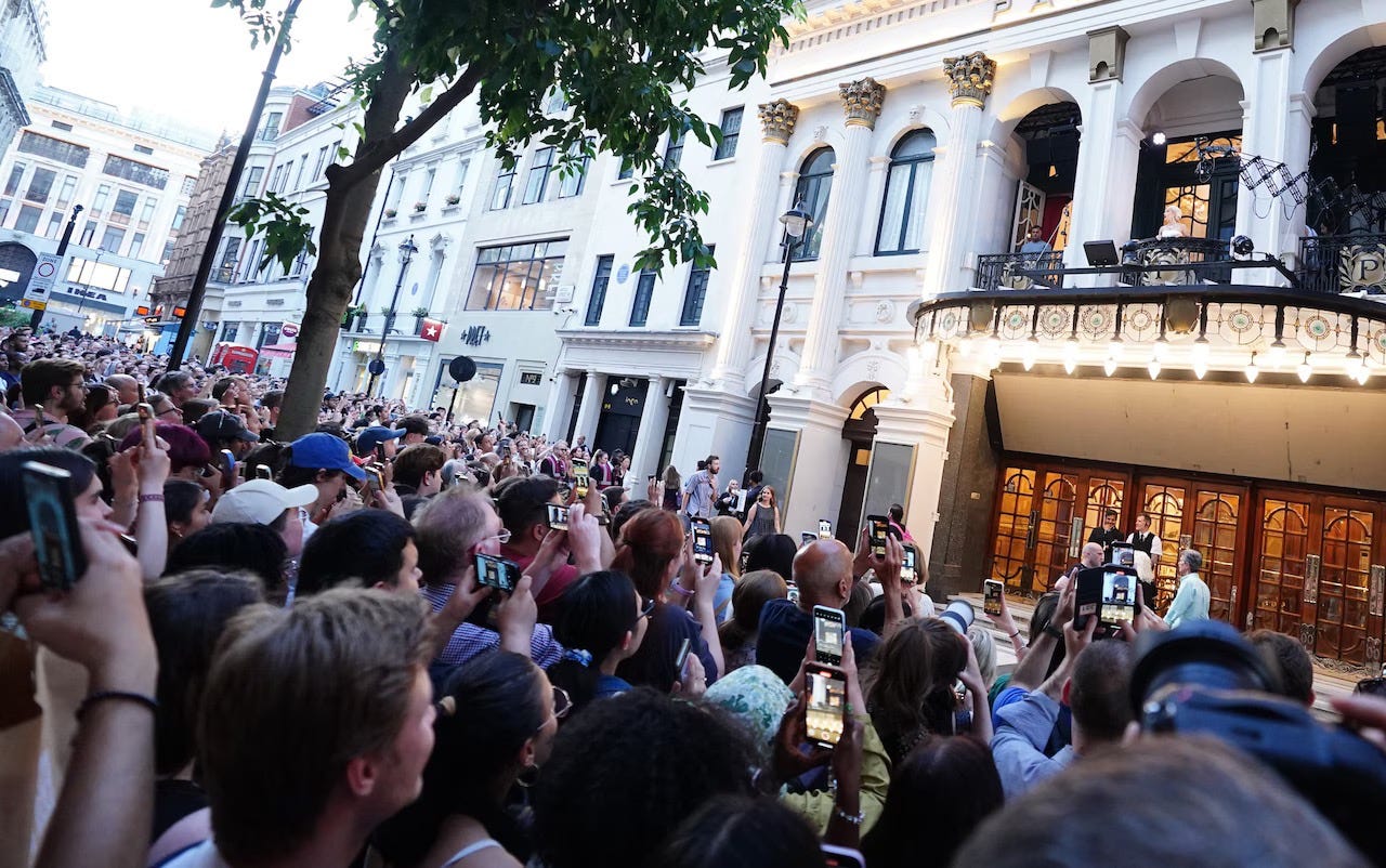 A crowd of people are gathered closely, phones raised and pointed at the balcony across the street where Rachel Zegler appeared to sing Don’t Cry For Me, Argentina, which is being livecast to attendees inside the theater.