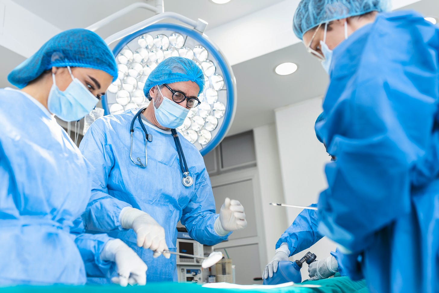 A group of surgeons in blue scrubs and surgical caps working in an operating room, with a large surgical light overhead and medical equipment in the background.