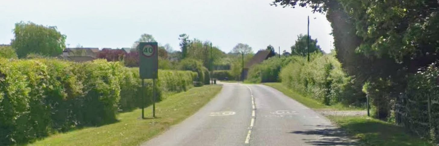 A single‑carriageway rural road in Great Chart marked with 40mph signs, bordered by hedges and trees, with no pavement, illustrating the area proposed for a new short footpath.