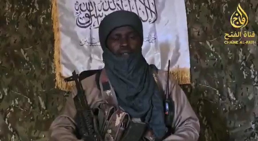 A man wearing a gray turban and camouflage uniform sits holding an assault rifle across his lap with a vest and equipment pouches. He has a serious expression and dark skin. Behind him is a green patterned wall with a large white flag bearing Arabic script and a gold emblem resembling a sword or emblem. Another similar flag hangs to the side. The text Chaine Al Fath appears in the lower right corner.