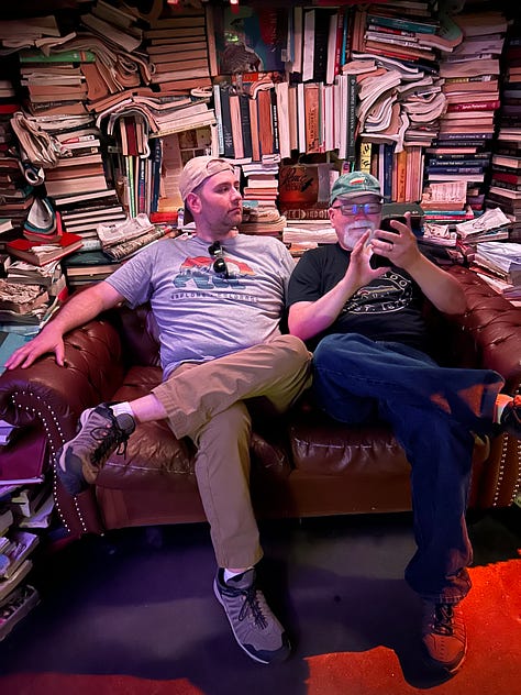 First picture shows a jumble of colorful items agains a green wall. One of the items is a trophy labeled Sports Ball. 2nd picture a young man and a man with a white beard sitting in front of two round windows. There is a green steel girder separating the sitters. #rd pic: The same two people are sitting on a couch in front of shelves with books piled haphazardly.