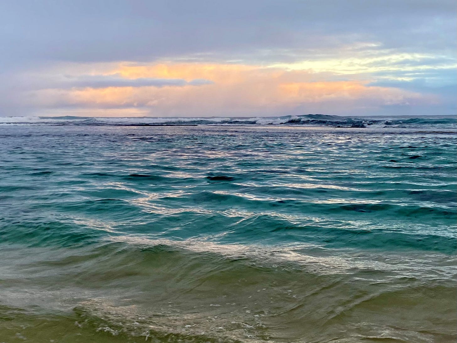 A picture of the ocean at sunrise. The sky is tinged pink. The water is smooth, deep teal 