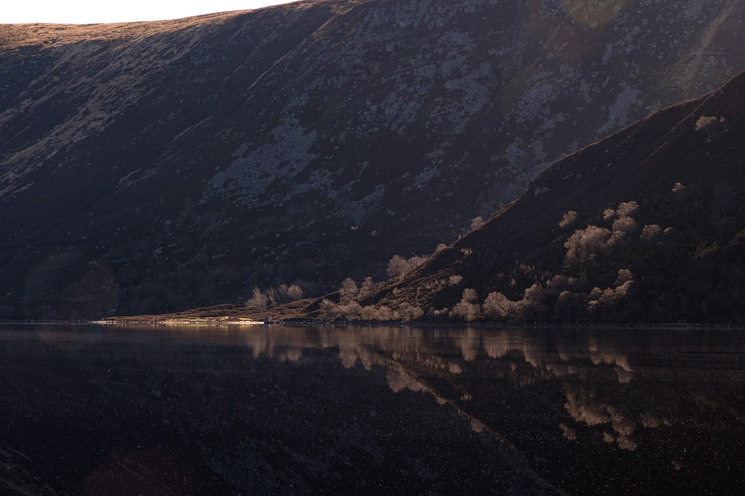 A moody photo of Loch Muick in the evening light. Parts of the surrounding hills are illuminated by the setting sun, while others remain in deep shadow. The loch is still and glass-like, with a dark, reflective surface. The scene feels calm, timeless, and suspended in the moment.