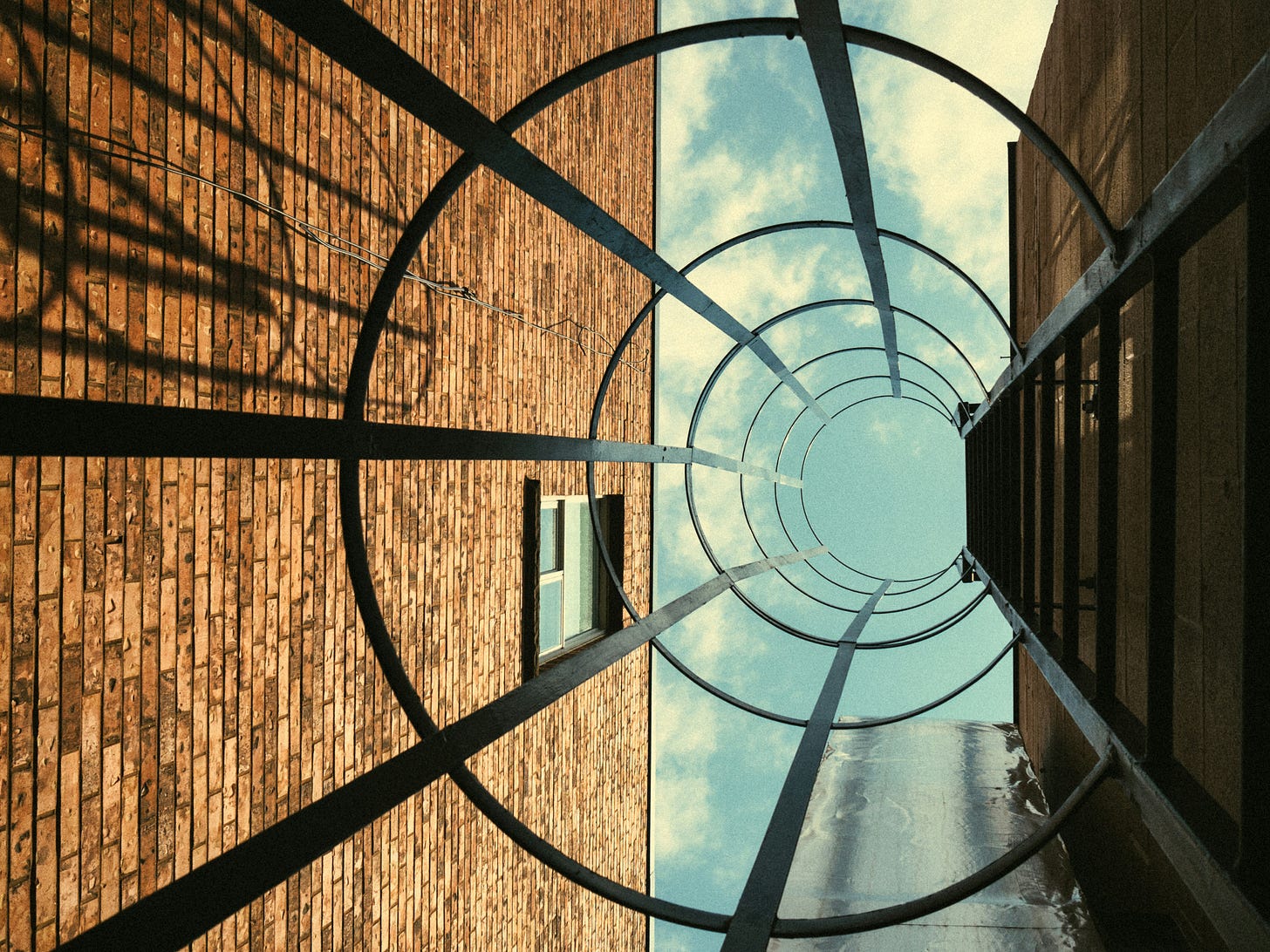 A worm's eye view of a caged ladder attached to the side of a brick building, looking up to a blue sky A worm's eye view of a caged ladder attached to the side of a brick building, looking up to a blue sky