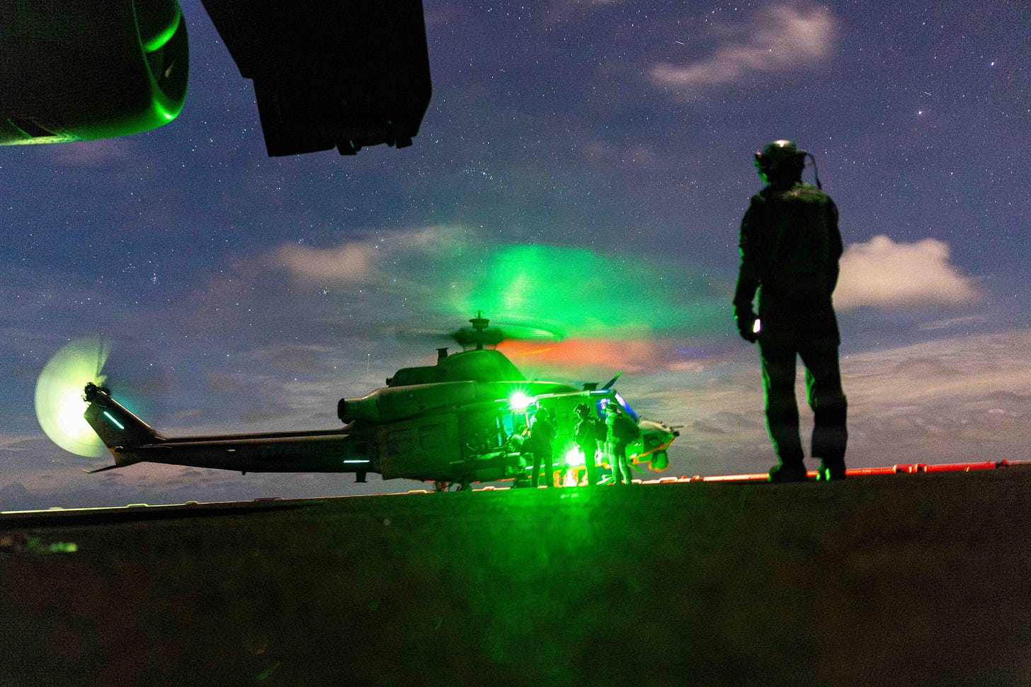 A Marine aircrewman silhouetted beside a helicopter lit green on a ship deck at night A Marine aircrewman silhouetted beside a helicopter lit green on a ship deck at night