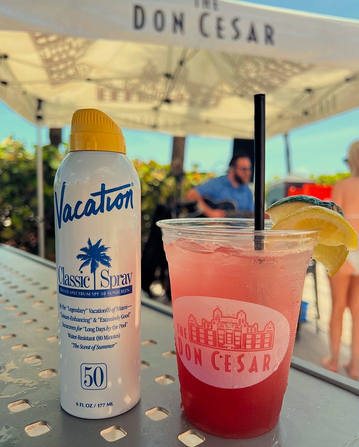 Beauty Editor wearing white-rimmed sunglasses on a sunny Florida beach with blue sky. And A bottle of Vacation Classic Spray SPF 50 sunscreen sitting on a table next to a pink tropical cocktail at The Don CeSar resort.