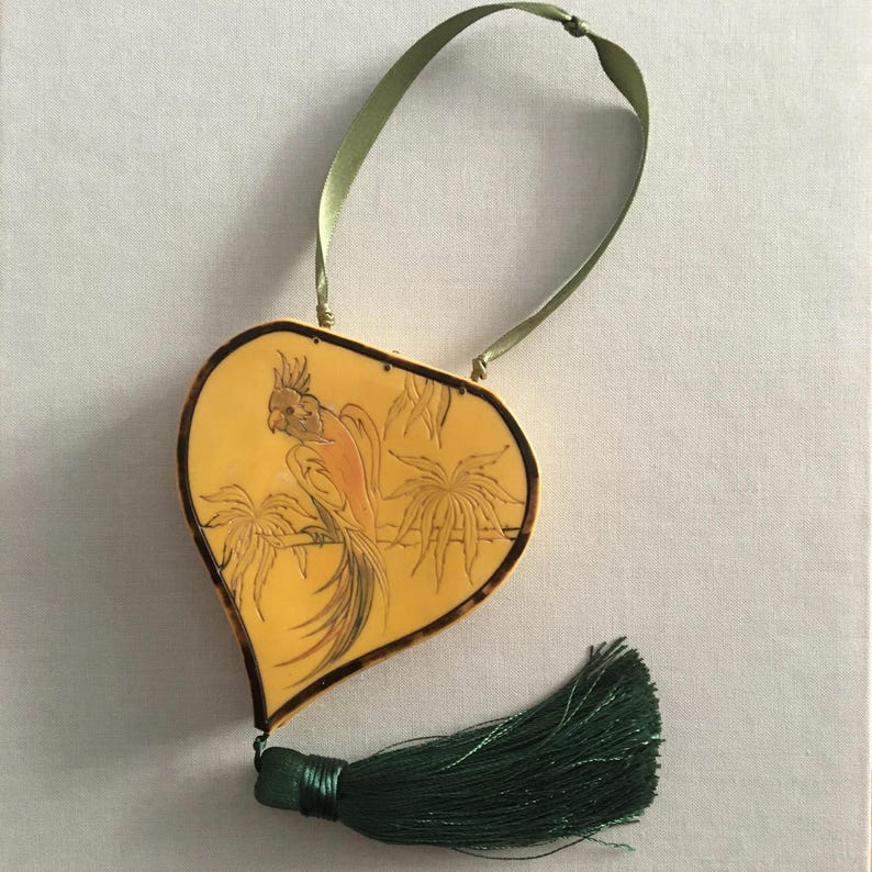 May include: A heart-shaped, yellow pendant with a parrot design and a green tassel. The pendant has a dark brown border and a green ribbon for hanging. The parrot is perched on a branch with palm trees in the background. The tassel is dark green.