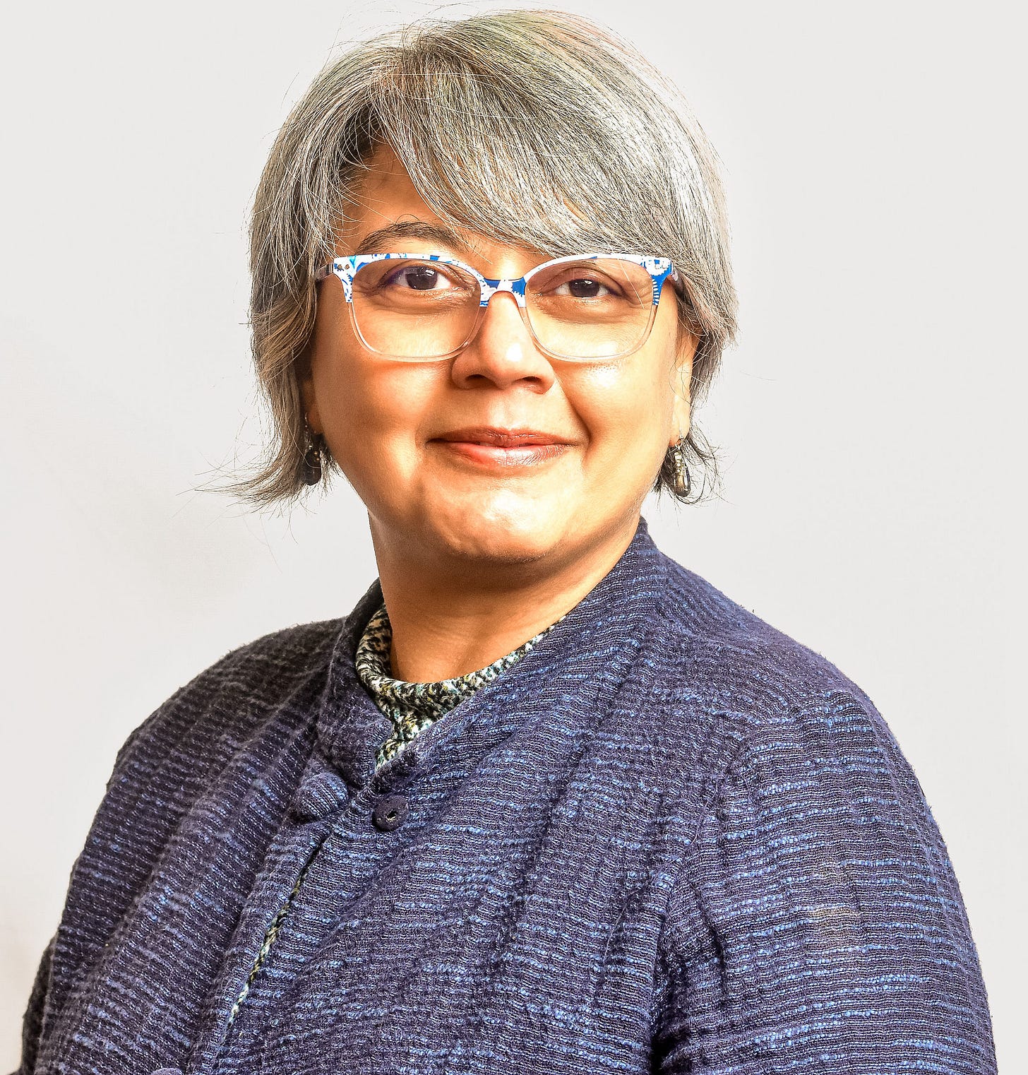 A studio-style head-and-shoulders portrait shows a person with short gray hair, patterned glasses, and small earrings, turned slightly toward the camera. They wear a textured blue jacket over a patterned top, with soft, even lighting that highlights facial features without harsh shadows. The neutral, light background and relaxed expression create a professional, calm, and approachable tone.