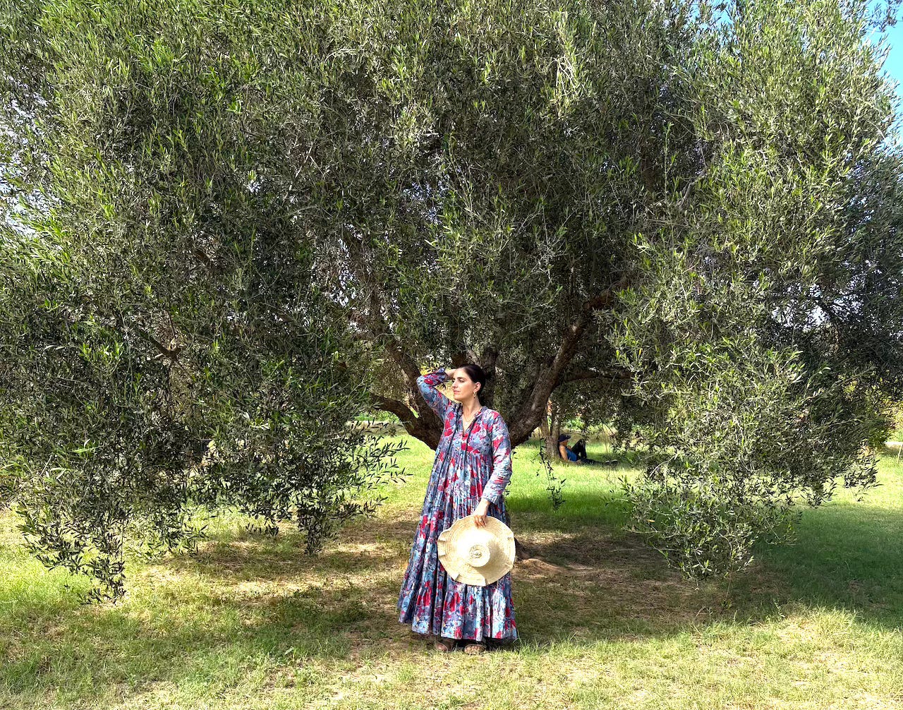 A woman in a colorful dress with a wide brimmed hat in her hand stands in the shade of an olive tree