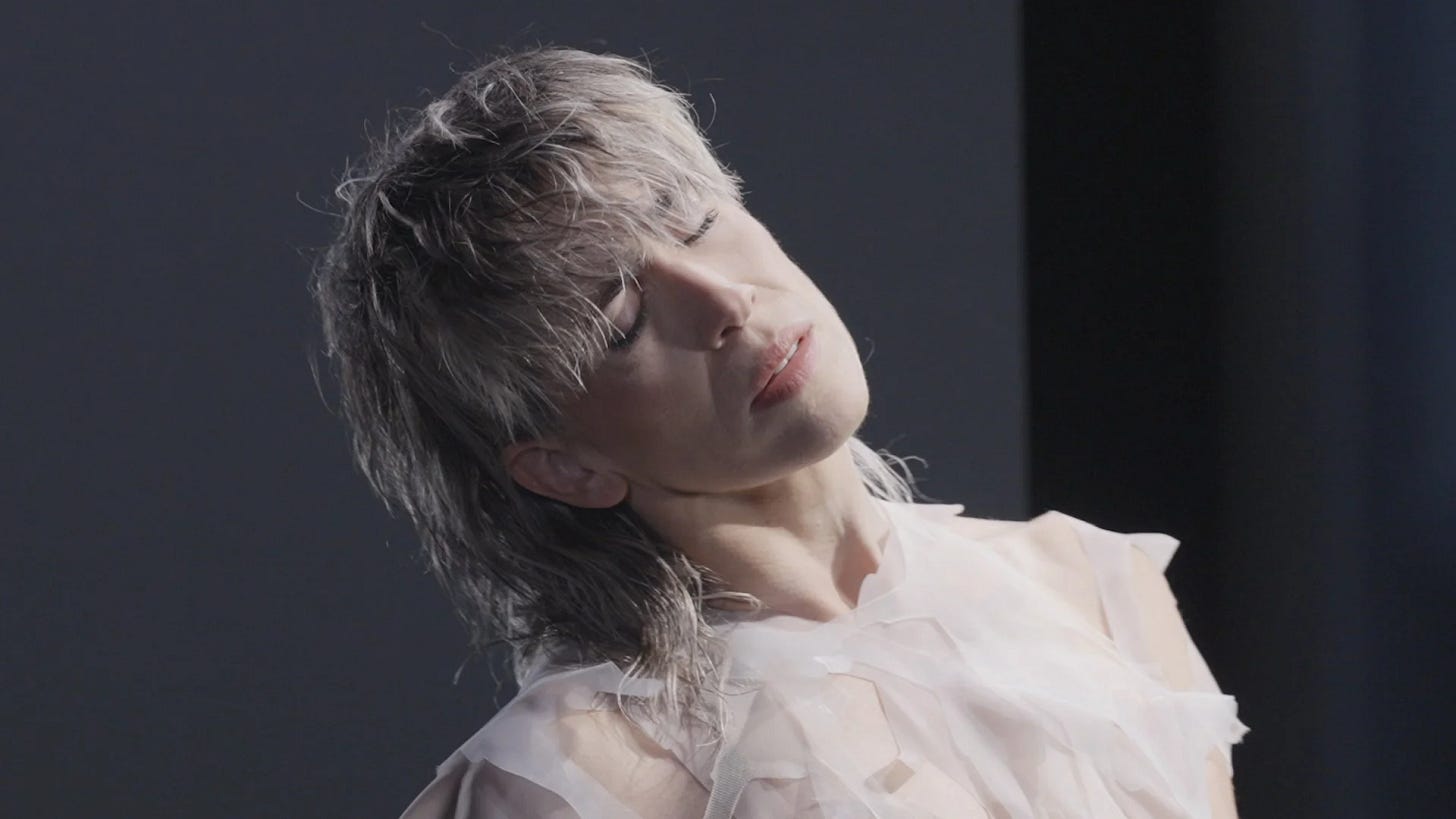A photo of a white person with cropped blonde hair in an indoor setting. They wear a white top and stand with their eyes closed, in a neutral pose. The background is dark.