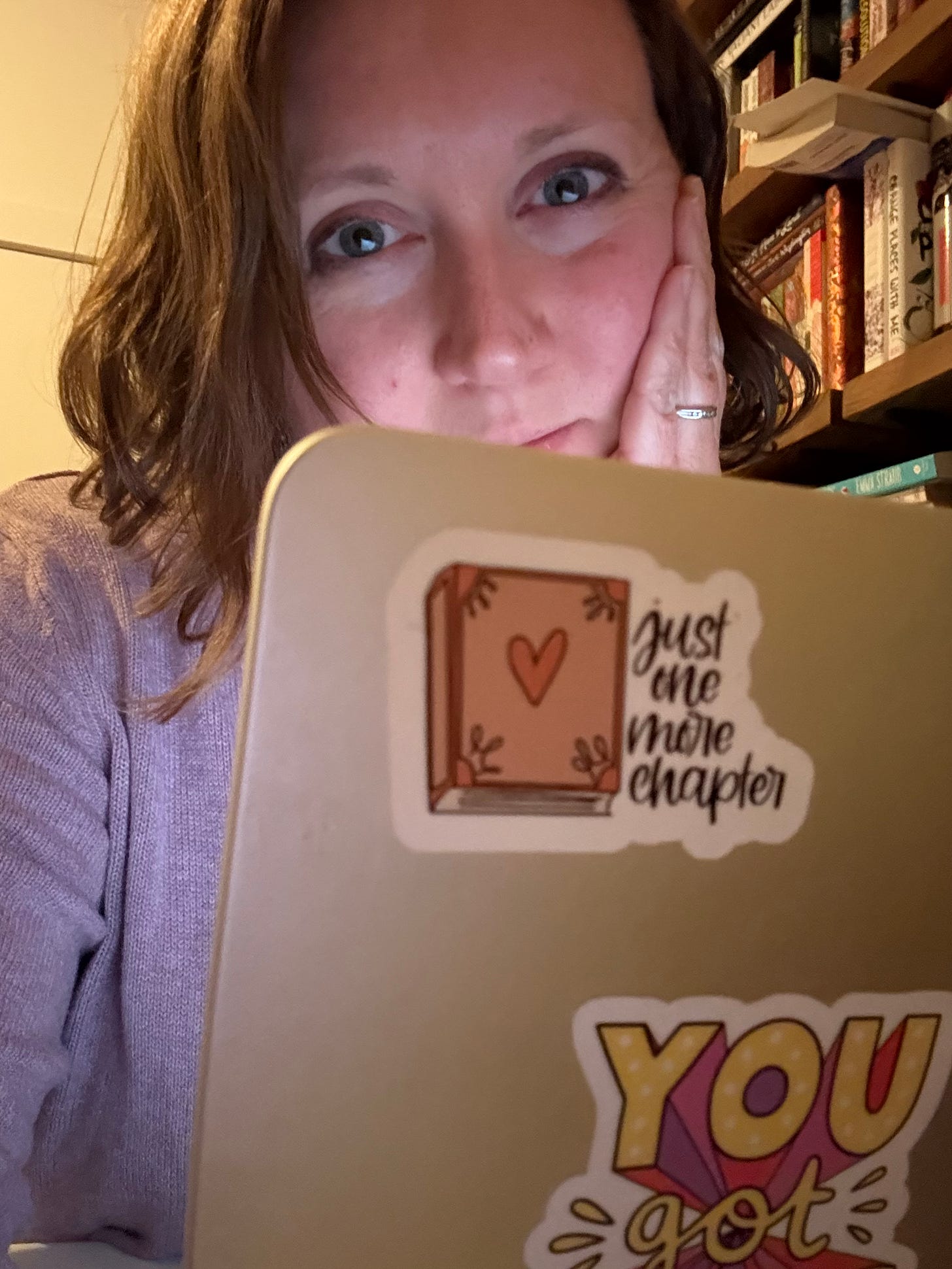 Kathryn, a white woman with brown hair and blue eyes, looks into the camera over the back of her laptop, which has a sticker saying Just One More Chapter. A bookshelf is in the background.
