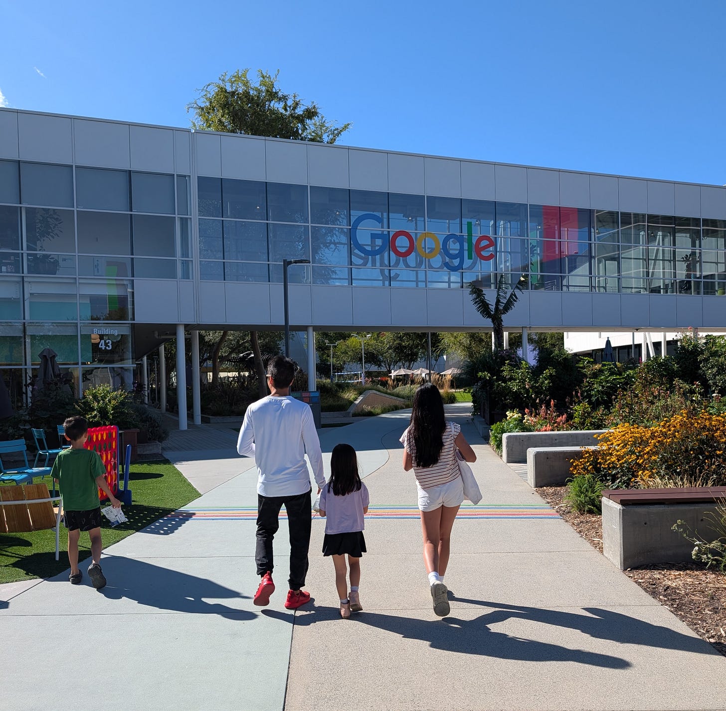 In the background is a building with the Google logo on it. In the foreground four people are walking towards the building, with their backs to the camera. There is an Asian man holding the hand of a little girl, an adolescent girl, and a little boy.