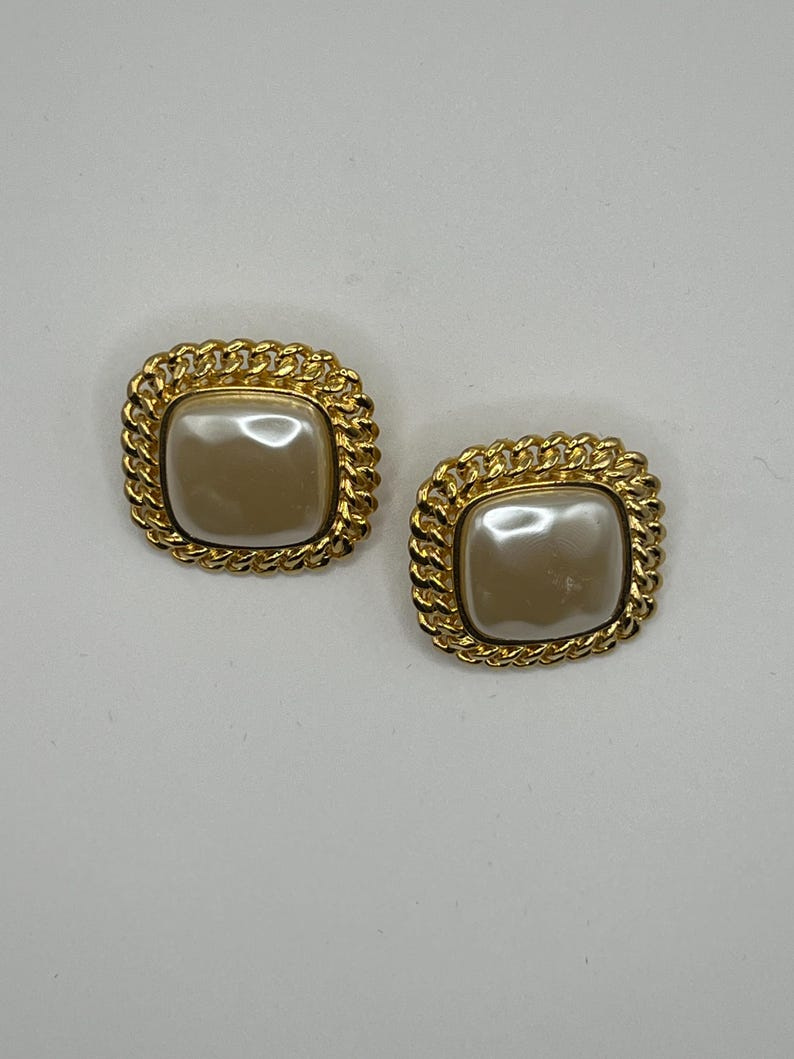 May include: Pair of gold-tone clip-on earrings. Each earring features a square-shaped, off-white faux pearl cabochon set within a gold-tone chain-link border. The earrings have a vintage or retro aesthetic, suitable for adding a touch of classic elegance to any outfit. The gold-tone metal is shiny and the faux pearls have a smooth, lustrous finish. These statement earrings are perfect for adding a touch of sophistication to a casual or formal look.