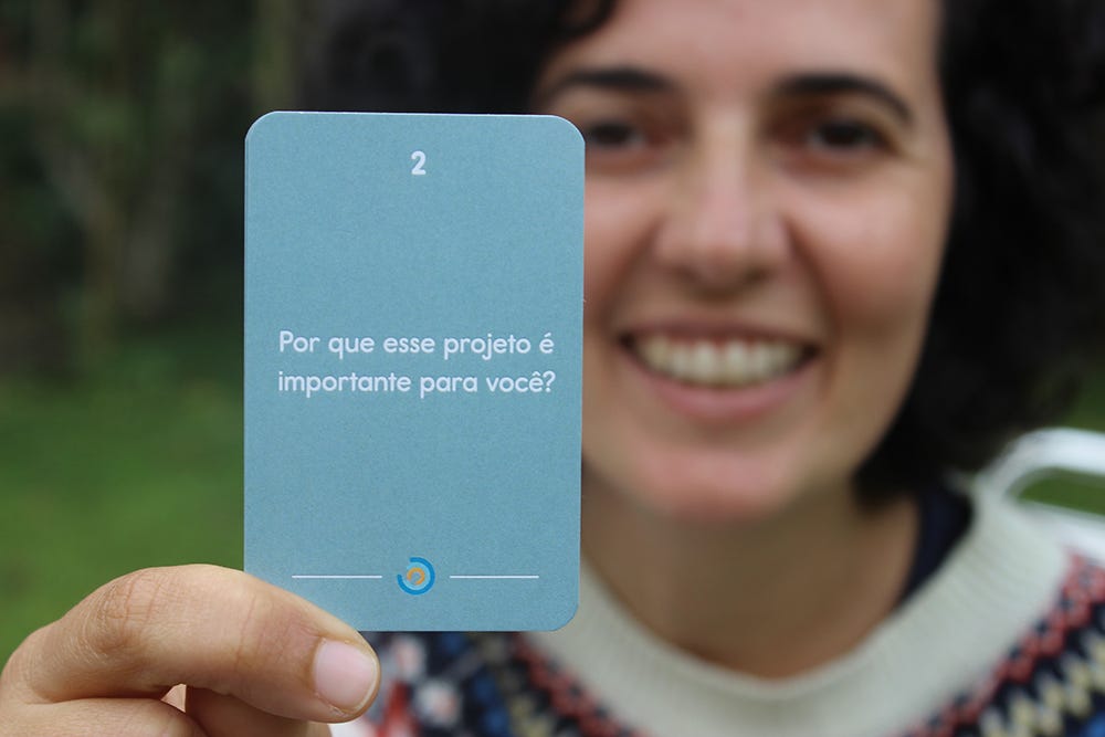 nessa foto a Mari aparece segurando uma carta do Combinados onde se lê "Por que esse projeto é importante para você?". Mari é uma mulher branca, de cabelos pretos e enrolados, e está vestindo uma blusa de lã branca com estampas azuis, amarelas e laranjas na gola. ela está sentada em uma cadeira de palha, num jardim com algumas árvores ao fundo.