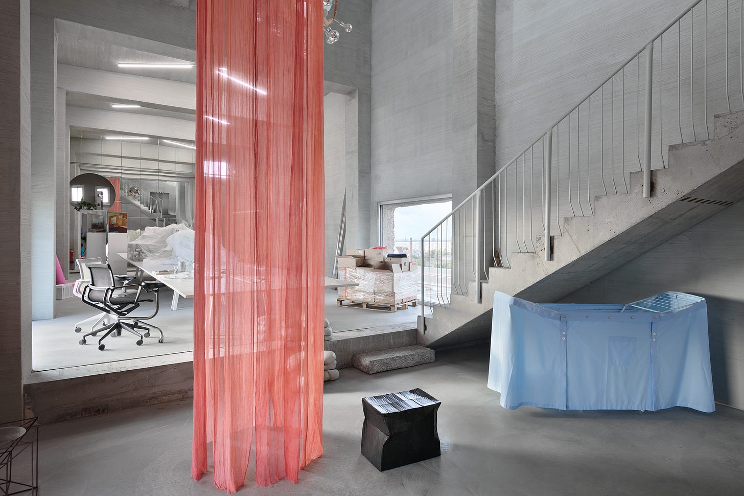 This is an interior photograph of a large, industrial-style room with rough gray walls and a concrete staircase leading up the right side. The scene is dominated by a long, sheer, shredded orange-red curtain hanging in the foreground, juxtaposed with a light blue fabric with shirt buttons covering a laundry rack and a glimpse into an adjacent, brighter work area.