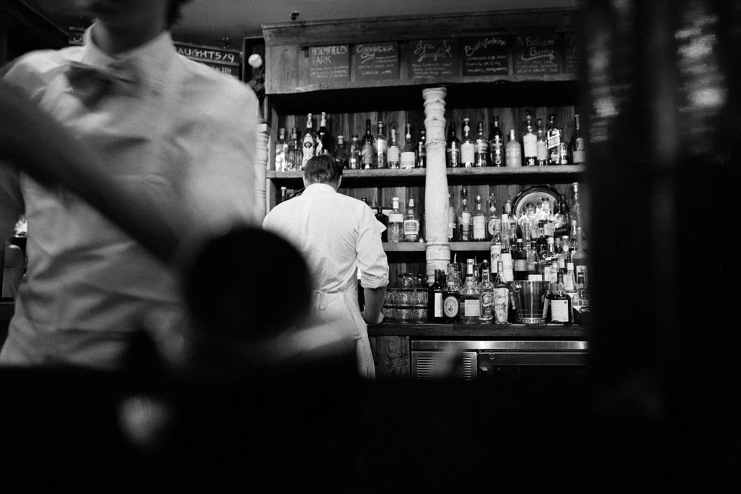 B&W photo of bar scene with bartender, waiter, and shelved alcohol. B&W photo of bar scene with bartender, waiter, and shelved alcohol.