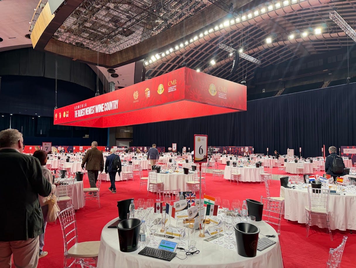 The Concours Mondial de Bruxelles judging hall in Yerevan, Armenia, with numbered tables set with wine glasses, spit buckets, and international flags beneath a banner reading 'Armenia: The Oldest Newest Wine Country